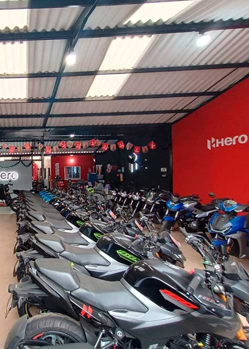 Generar una imagen fotorrealista de la fachada del concesionario Mundo Hero, basada estrictamente en la imagen de referencia que se adjunta. No modificar la infraestructura, arquitectura, colores, proporciones ni distribución del local. Mantener exactamente la misma fachada, ángulos y estructura visibles. Integrar el logo oficial de Mundo Hero (adjuntado) de forma natural y realista en la fachada, respetando su diseño original, colores, proporciones y tipografía, sin alteraciones. La imagen debe transmitir un ambiente profesional, moderno y confiable, con iluminación equilibrada (día claro), sombras realistas y alto nivel de detalle. Estilo fotográfico realista, calidad alta, enfoque nítido, sin personas ficticias, sin elementos añadidos que no existan en la imagen original. El objetivo es obtener una versión visualmente mejorada y limpia de la fachada real, no una reinterpretación creativa.